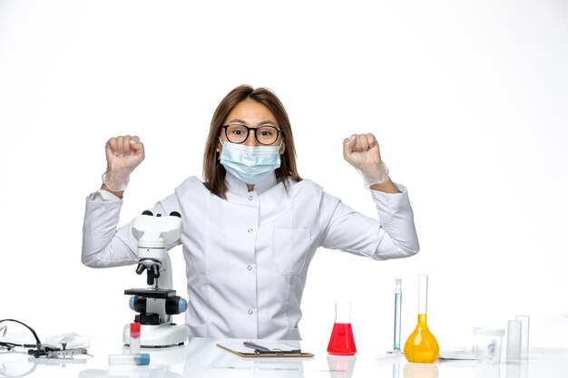 Vista frontal doctora en traje médico blanco y máscara debido a covid usando microscopio en escritorio blanco