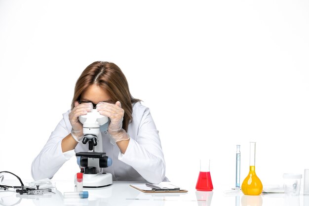 Vista frontal doctora en traje médico blanco y máscara debido a covid usando microscopio en espacio en blanco