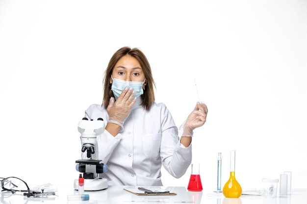 Vista frontal doctora en traje médico blanco y máscara debido a covid usando microscopio en espacio en blanco