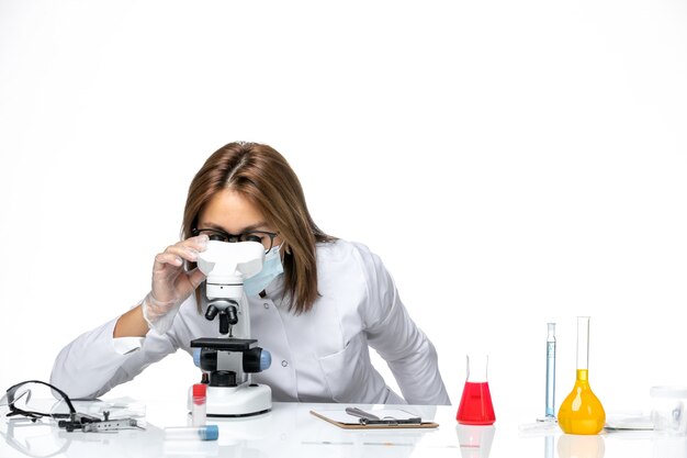 Foto gratuita vista frontal doctora en traje médico blanco y máscara debido a covid usando microscopio en espacio en blanco