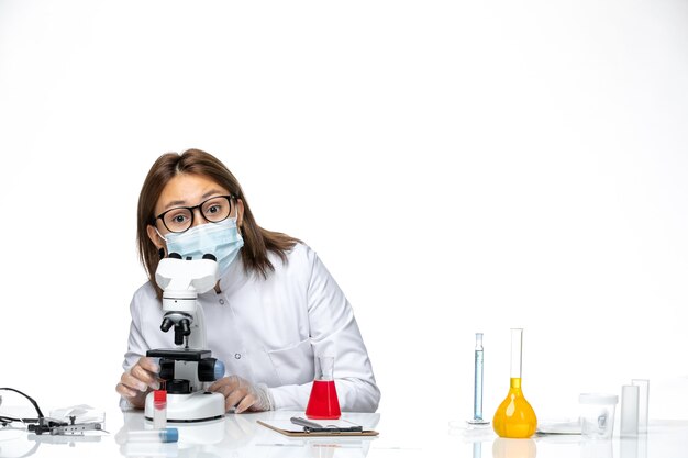 Vista frontal doctora en traje médico blanco con máscara debido a covid usando microscopio en espacio en blanco