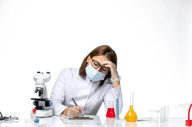 Vista frontal doctora en traje médico blanco con máscara debido a que covid escribe algo en un espacio en blanco claro