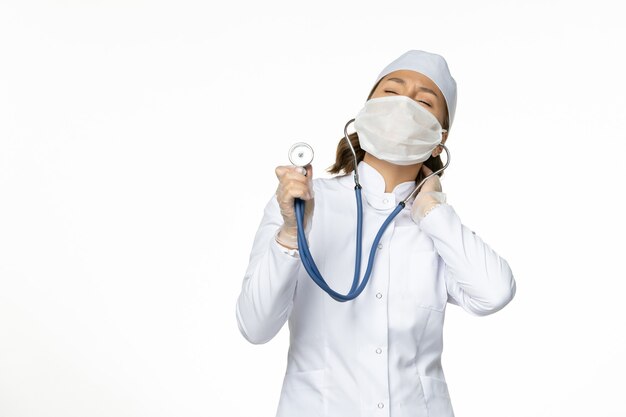 Vista frontal doctora en traje médico blanco y máscara con estetoscopio en el aislamiento del virus de la medicina de la enfermedad pandémica de escritorio blanco