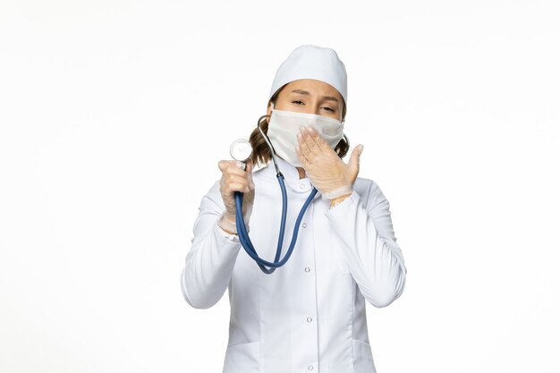 Vista frontal doctora en traje médico blanco y máscara con estetoscopio bostezando en la pared blanca medicina de enfermedad de virus pandémico