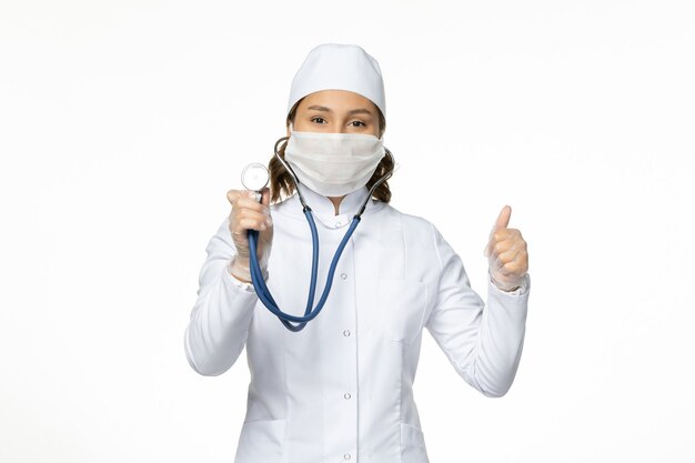 Vista frontal doctora en traje médico blanco y máscara con estetoscopio en la pared blanca medicina de la enfermedad del virus de la pandemia