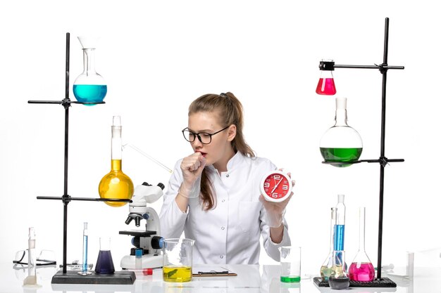 Vista frontal doctora en traje médico blanco sosteniendo relojes y tosiendo sobre fondo blanco química del virus covid pandémico