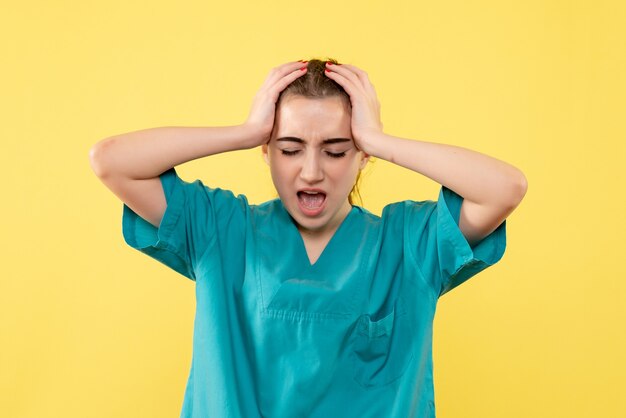 Vista frontal de la doctora en traje médico con dolor de cabeza en la pared amarilla