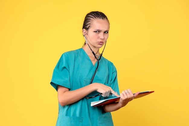Vista frontal de la doctora en traje médico con estetoscopio y análisis en pared amarilla