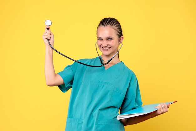 Vista frontal de la doctora en traje médico con estetoscopio y análisis en pared amarilla