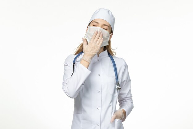 Vista frontal doctora en traje médico con máscara debido al coronavirus que bosteza en la pared blanca enfermedad covid-enfermedad pandémica del virus