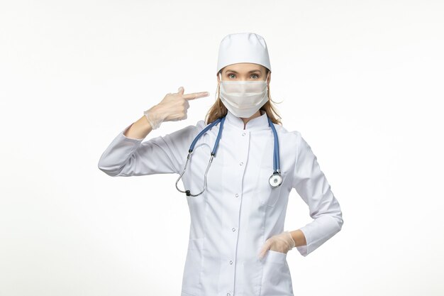 Vista frontal doctora en traje médico con máscara debido a coronavirus en la enfermedad de la pared blanca enfermedad covid-enfermedad del virus pandémico