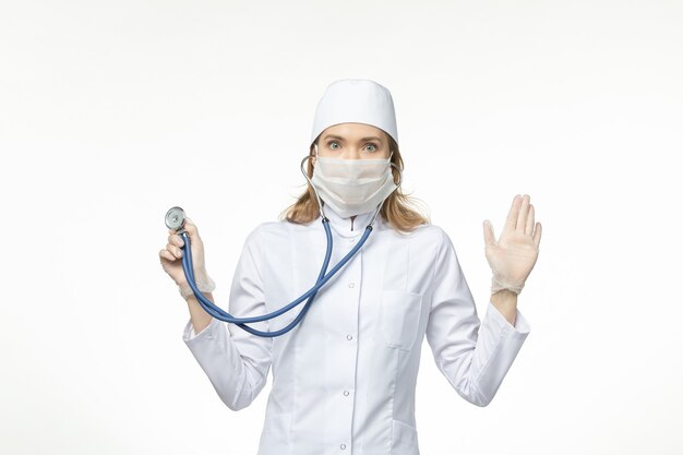Vista frontal doctora en traje médico con máscara debido a coronavirus sosteniendo estetoscopio en la pared blanca clara virus pandemia enfermedad covid