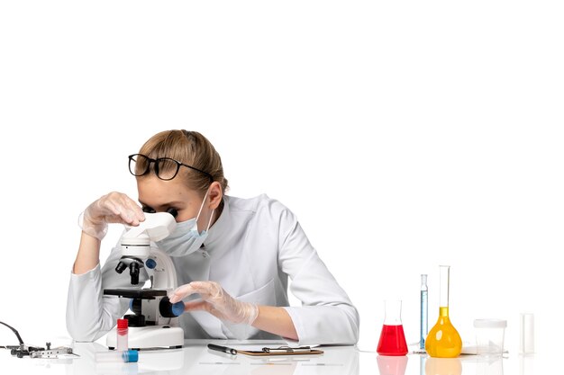 Vista frontal doctora en traje médico con máscara debido a covid usando microscopio en un espacio en blanco claro