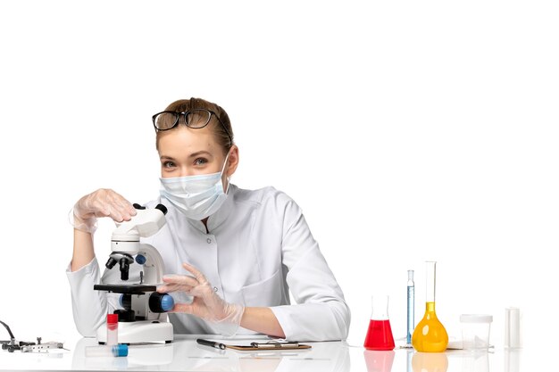 Vista frontal doctora en traje médico con máscara debido a covid usando microscopio en un espacio en blanco claro