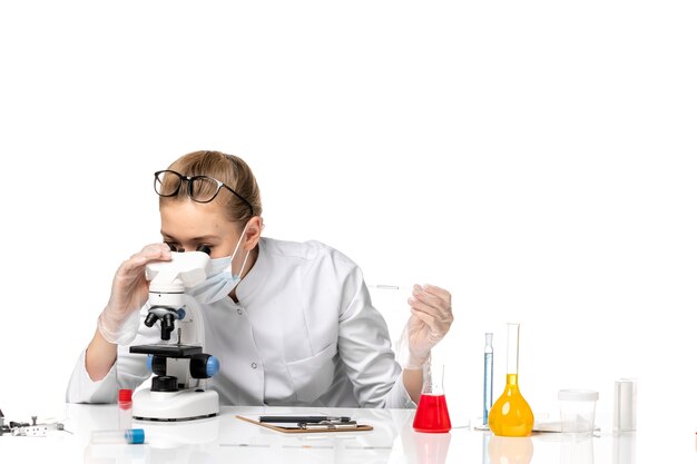 Vista frontal doctora en traje médico con máscara debido a covid usando microscopio en el espacio en blanco