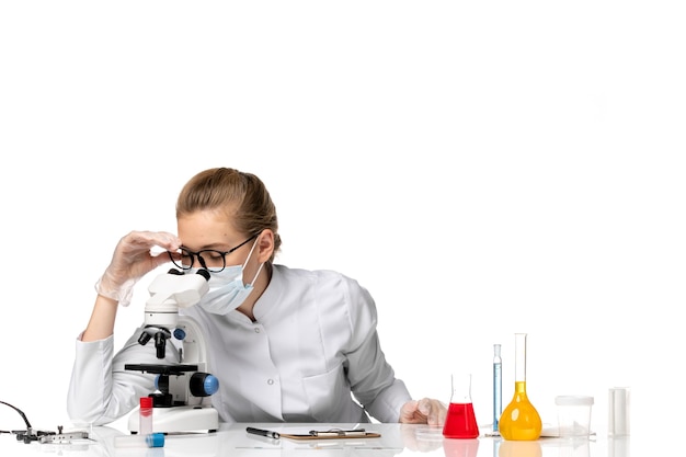 Vista frontal doctora en traje médico con máscara debido a covid usando microscopio en el espacio en blanco