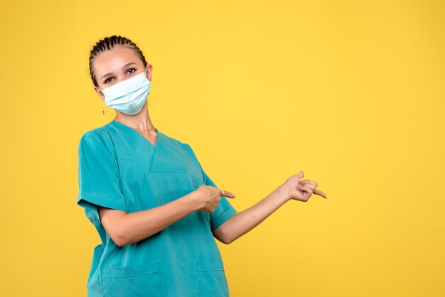 Vista frontal de la doctora en traje médico y máscara estéril en una pared amarilla