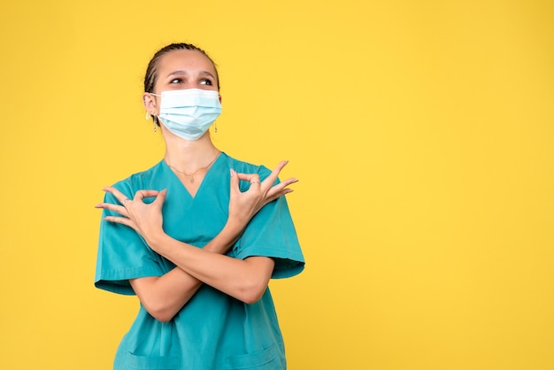 Vista frontal de la doctora en traje médico y máscara estéril en una pared amarilla