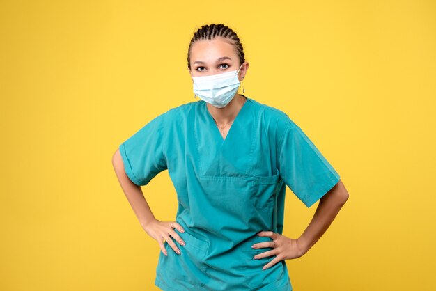 Vista frontal de la doctora en traje médico y máscara estéril en una pared amarilla