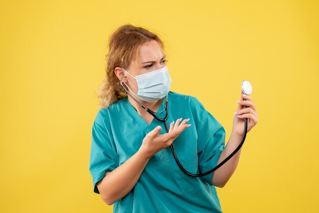 Vista frontal de la doctora en traje médico y máscara con estetoscopio en pared amarilla