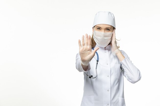 Vista frontal doctora en traje médico con máscara y guantes debido al coronavirus en la pared blanca clara enfermedad covid-virus pandémico