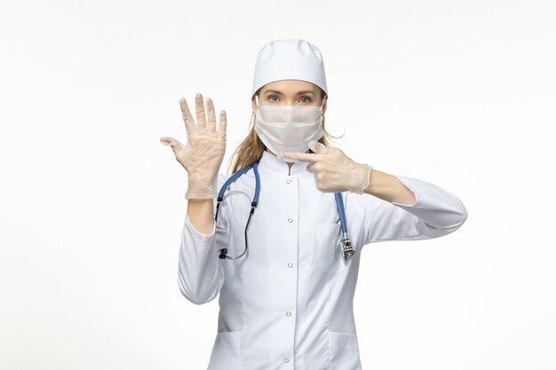 Vista frontal doctora en traje médico con máscara y guantes debido al coronavirus en la pared blanca enfermedad covid-enfermedad pandémica del virus