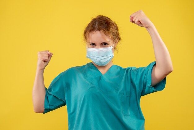 Vista frontal de la doctora en traje médico y máscara regocijándose en la pared amarilla