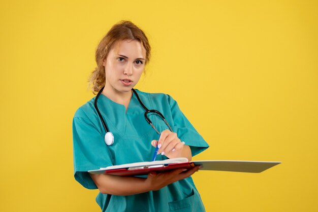 Vista frontal de la doctora en traje médico con papeles y análisis en la pared amarilla