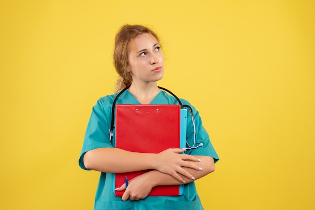 Vista frontal de la doctora en traje médico con papeles y análisis en la pared amarilla