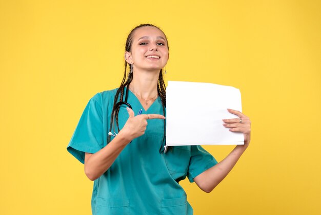 Vista frontal de la doctora en traje médico sosteniendo papeles en la pared amarilla