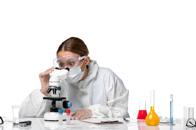 Vista frontal doctora en traje de protección y con máscara con microscopio sobre fondo blanco coronavirus pandémico de virus covid de salud