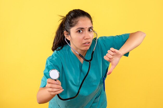 Vista frontal de la doctora en uniforme sosteniendo estetoscopio haciendo pulgar hacia abajo firmar en la pared amarilla