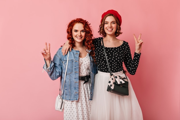 Vista frontal de dos amigos abrazados sobre fondo rosa. Foto de estudio de niñas sonrientes mostrando signos de paz.