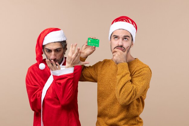 Vista frontal de dos amigos uno con abrigo de santa mirando su mano y el otro sosteniendo una tarjeta sobre fondo beige aislado