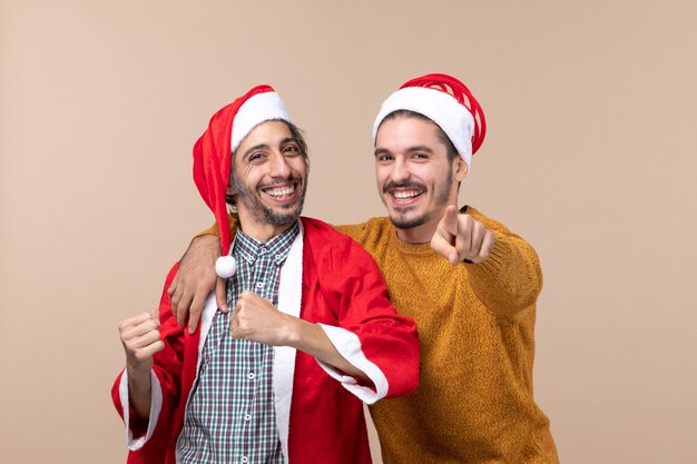 Vista frontal de dos amigos con gorro de Papá Noel mirando a la cámara sobre fondo beige aislado