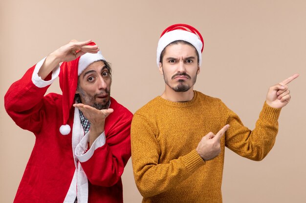 Vista frontal de dos amigos con gorro de Papá Noel que muestra la dirección con cara seria sobre fondo beige aislado