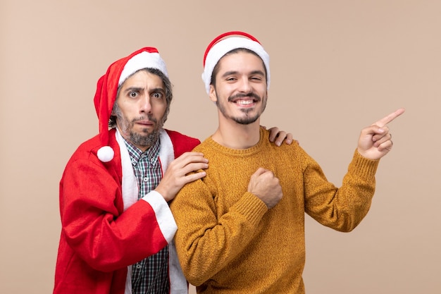 Vista frontal de dos amigos con gorro de Papá Noel, uno sosteniendo el hombro de amigos y el otro mostrando la dirección sobre fondo beige aislado