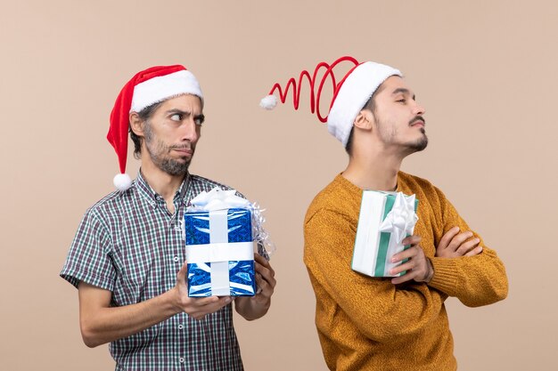 Vista frontal de dos amigos ofendidos con gorro de Papá Noel y sosteniendo regalos de Navidad sobre fondo beige aislado