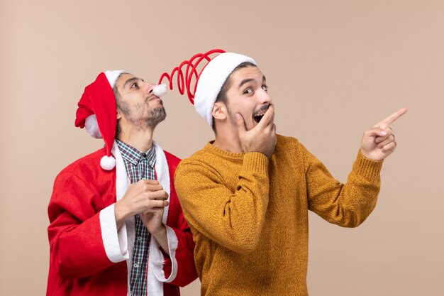 Vista frontal de dos amigos con sombreros de santa, uno tocando sombreros pompón y el otro mostrando la dirección sobre fondo beige aislado