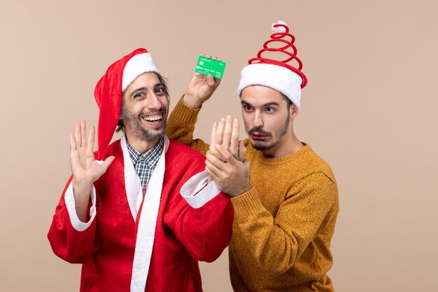 Vista frontal de dos chicos, uno con abrigo de santa riendo y el otro con tarjeta sobre fondo beige aislado