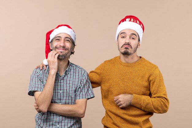 Vista frontal de dos chicos confundidos con gorro de Papá Noel, uno poniendo su mano sobre el hombro de los demás sobre fondo beige aislado