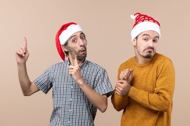 Vista frontal de dos chicos confundidos con sombreros de santa un dedo que muestra algo sobre fondo beige aislado