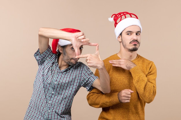 Vista frontal de dos chicos curiosos con gorro de Papá Noel uno haciendo signo de cámara con sus manos sobre fondo aislado