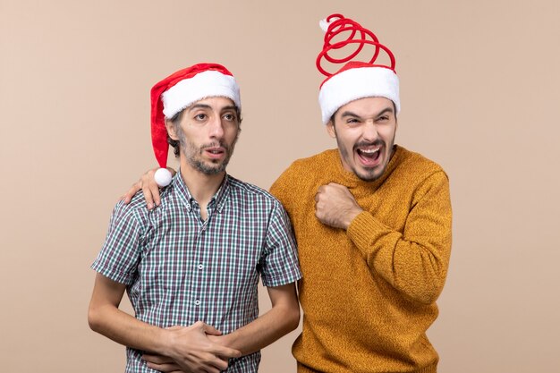 Vista frontal de dos chicos un deprimido y feliz con gorro de Papá Noel uno poniendo su mano sobre el hombro del otro sobre fondo beige aislado