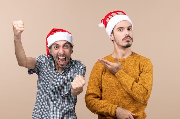 Vista frontal dos chicos un eufórico y un confundido con gorro de Papá Noel, uno mostrando el otro sobre fondo beige aislado