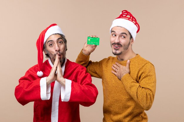 Vista frontal de dos chicos felices, uno con abrigo de santa y el otro con crédito poniendo su mano sobre su pecho sobre fondo beige aislado