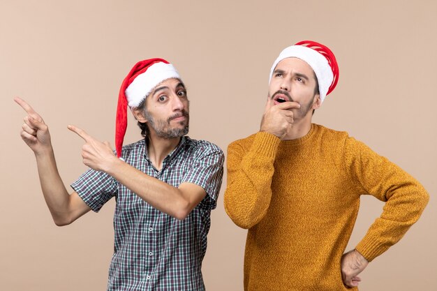 Vista frontal de dos chicos con gorro de Papá Noel, uno mostrando algo y el otro pensando y llevándose la mano a la boca sobre fondo beige aislado