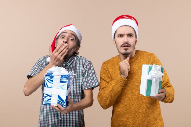 Vista frontal de dos chicos maravillados con gorro de Papá Noel y sosteniendo presenta uno poniendo la mano en la boca sobre fondo beige aislado
