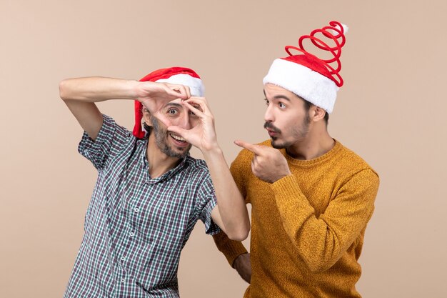 Vista frontal de dos chicos de Navidad con gorro de Papá Noel, uno haciendo forma de corazón con sus manos sobre fondo beige aislado