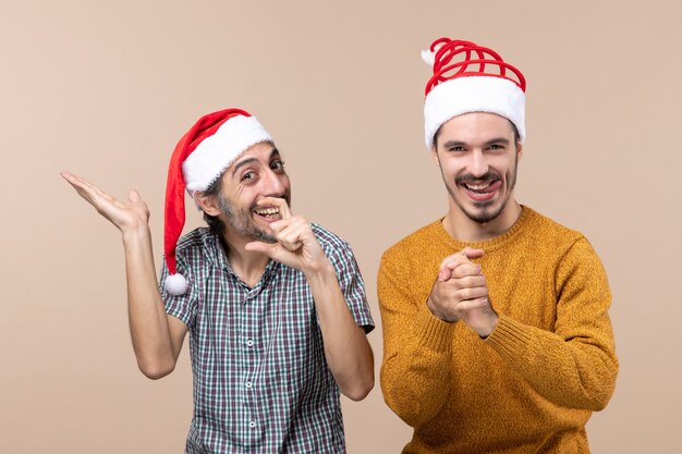 Vista frontal de dos chicos riendo con gorro de Papá Noel, uno mostrando algo y el otro sosteniendo sus manos juntas sobre fondo beige aislado